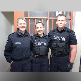 Two female and one male cadets, in uniform, smiling, standing in front of police jail cell
